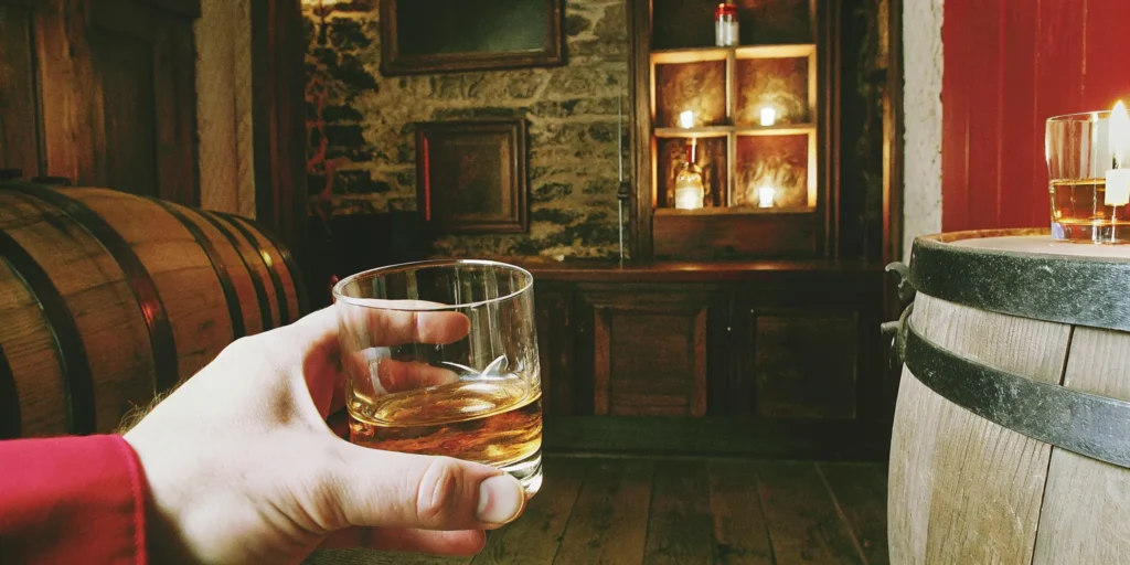 A hand holds a glass of whiskey in a bar, a guide for how to drink whiskey for beginners.