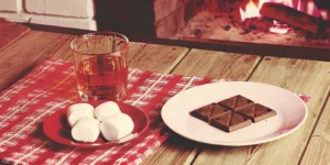 A glass of Mash and Mallow whiskey with marshmallows and chocolate on a rustic table by a fire.