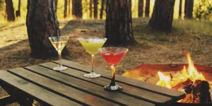 Easy cocktails to make while camping, served on a wooden table next to a glowing campfire.