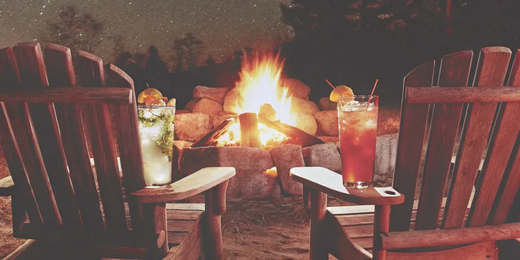 Two of the best campfire cocktails on Adirondack chairs next to a glowing fire.
