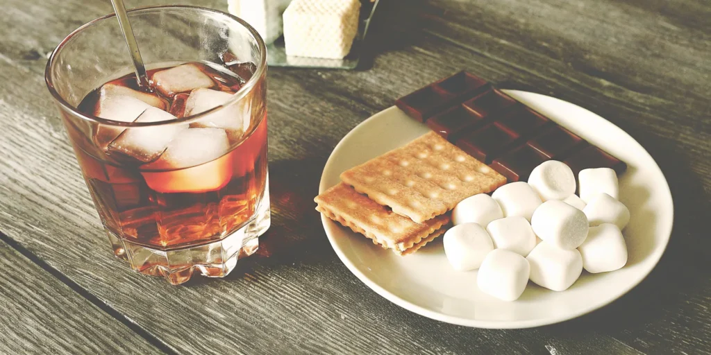 A glass of the best s'mores whiskey with chocolate, marshmallows, and graham crackers.