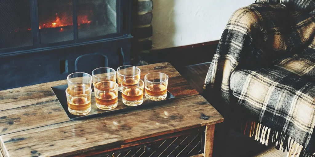 Six glasses of good starting whiskey on a rustic table near a fireplace.