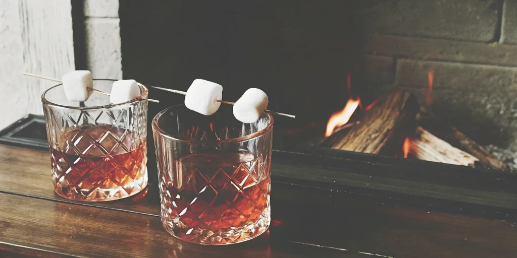 A Mash & Mallow gift set with two glasses of s'mores whiskey and toasted marshmallows by a cozy fireplace.