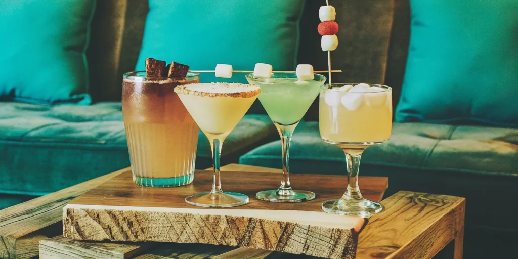 Smoky campfire cocktails with toasted marshmallow and chocolate garnishes on a wooden tray.