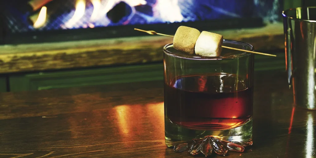 A Mash & Mallow old fashioned garnished with toasted marshmallows by a cozy fireplace.