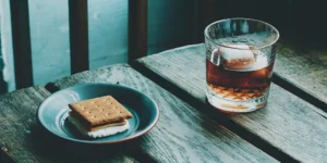 A glass of graham cracker flavored liquor on the rocks next to a plate of s'mores.