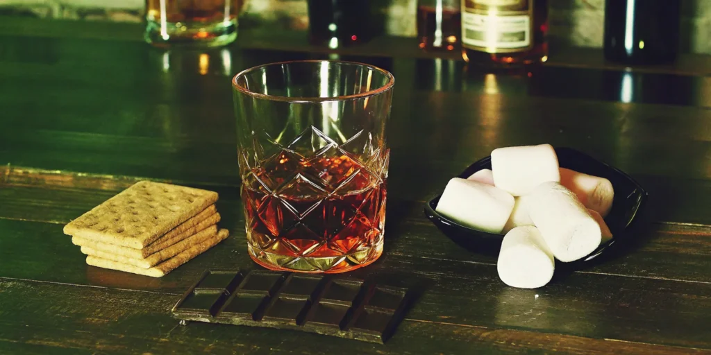 A glass of Mash and Mallow s'mores whiskey with graham crackers, chocolate, and marshmallows.