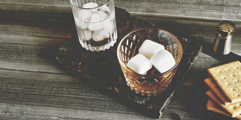 A glass of s'mores whiskey on the rocks with marshmallows and graham crackers.